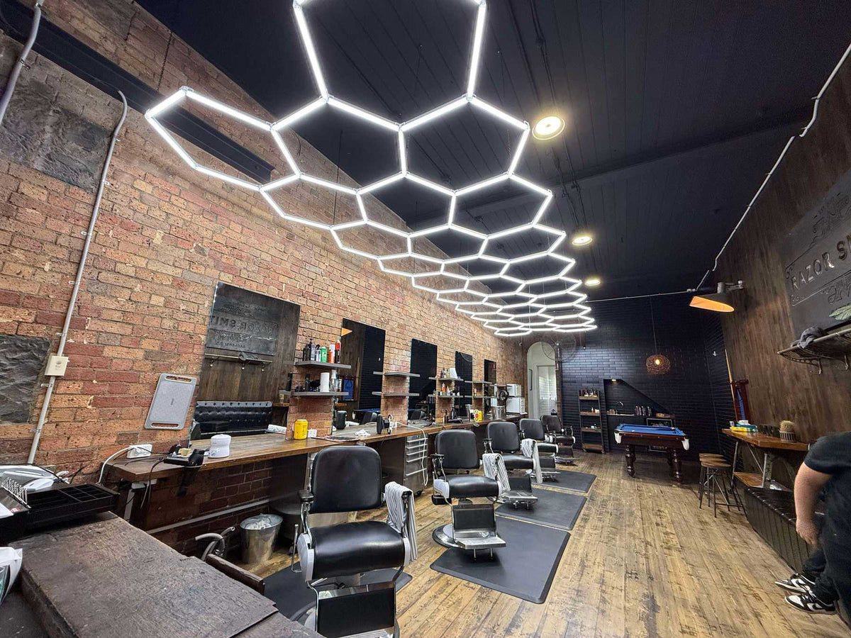 Barber chairs and mirrors in a brick-wall barber shop, under a long hexagon LED grid on a black ceiling