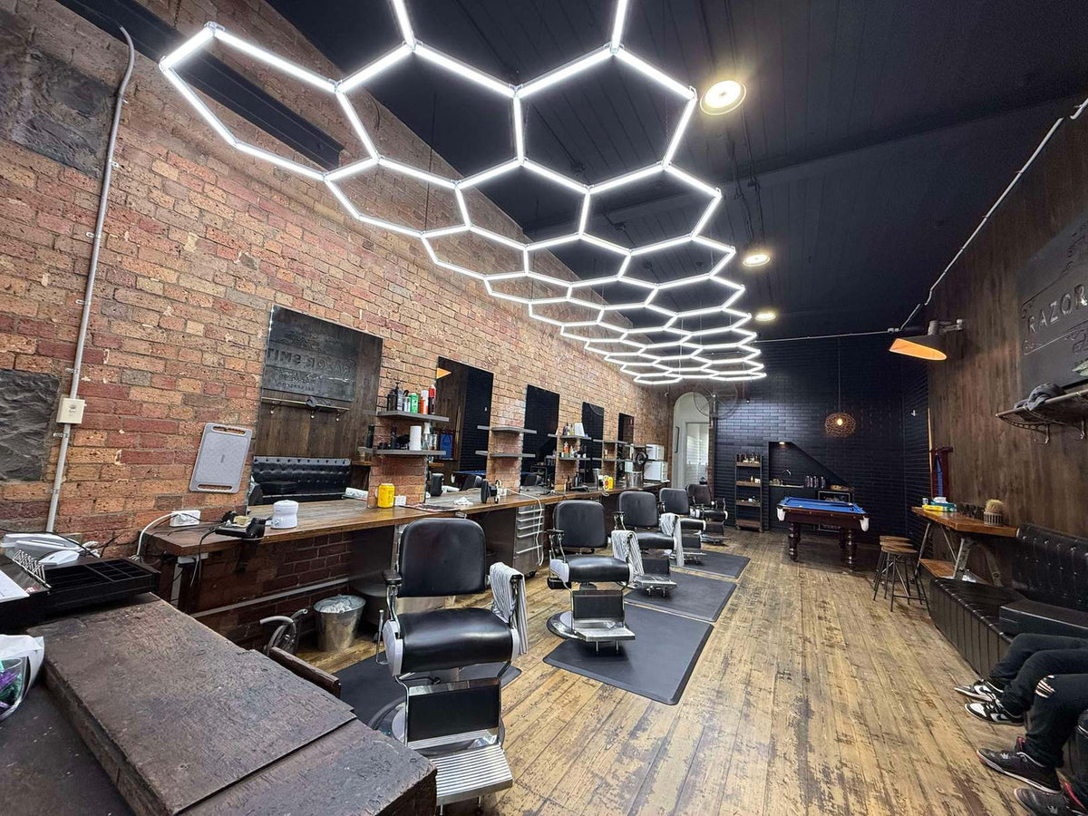 Long brick-wall barber shop with a hexagon LED grid running the length of the black ceiling above a row of barber chairs