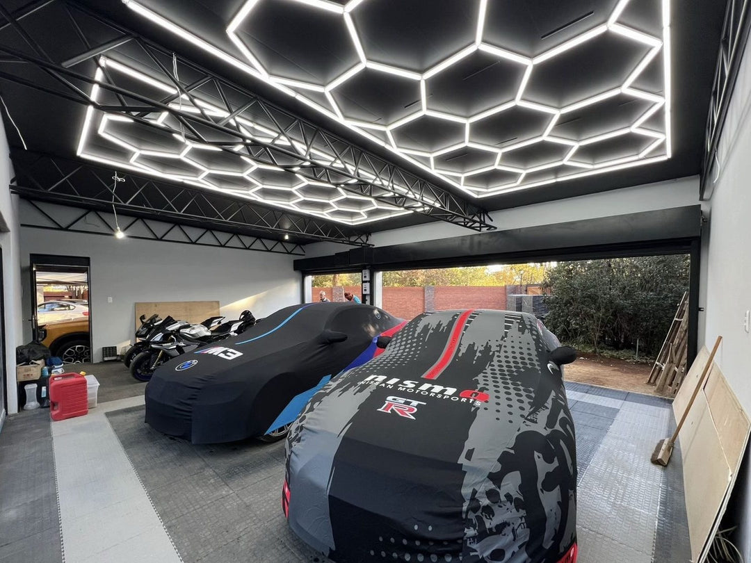 Two large hexagon LED grids under a black steel-beam garage ceiling above covered sports cars