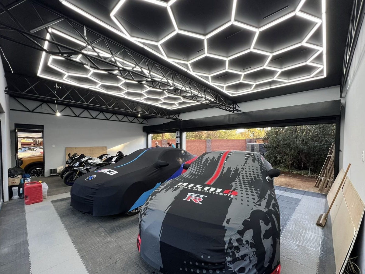Two large hexagon LED grids under a black steel-beam garage ceiling above covered sports cars