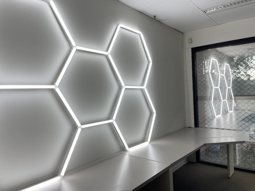 Large hexagon LED lights mounted on a white office wall above a long desk, with a second set visible through the window